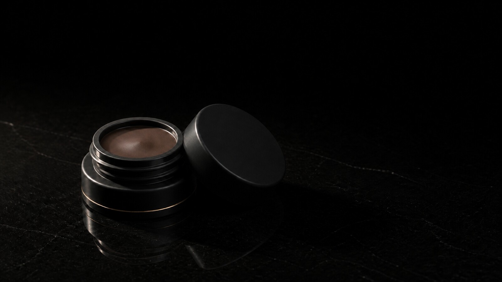 Brow Pomade — seen in three-quarter view, low in a wide frame — under a single warm gold light on black.