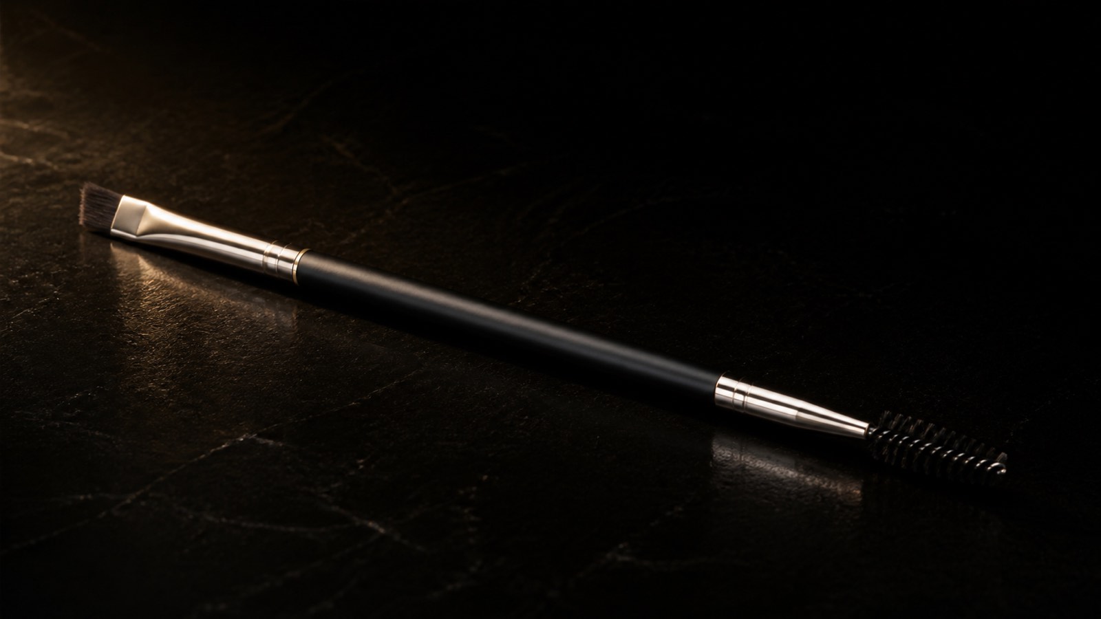 Brow Duo Brush — seen in three-quarter view, low in a wide frame — under a single warm gold light on black.