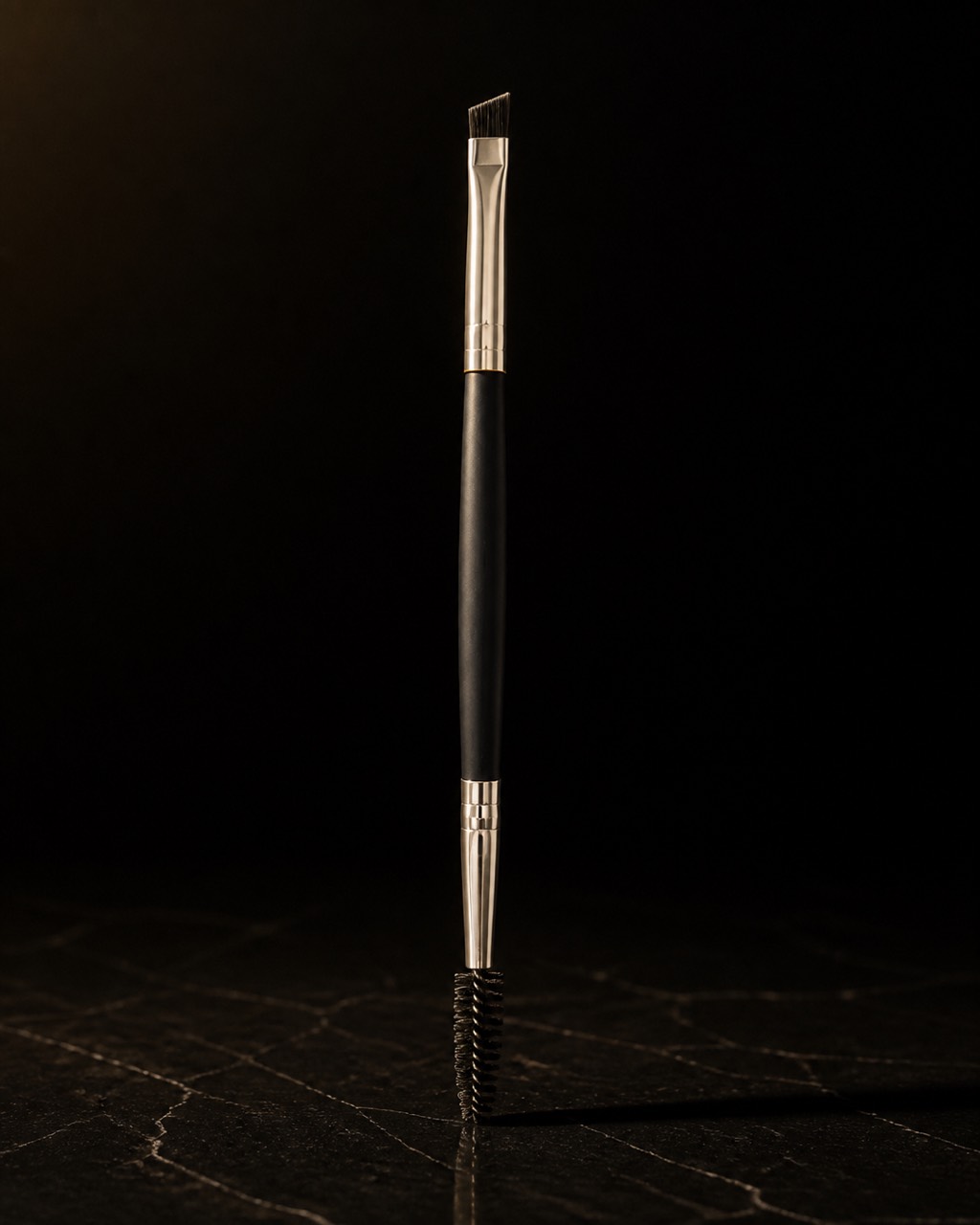 Brow Duo Brush — brow duo — standing alone against near-black under a single warm gold light.
