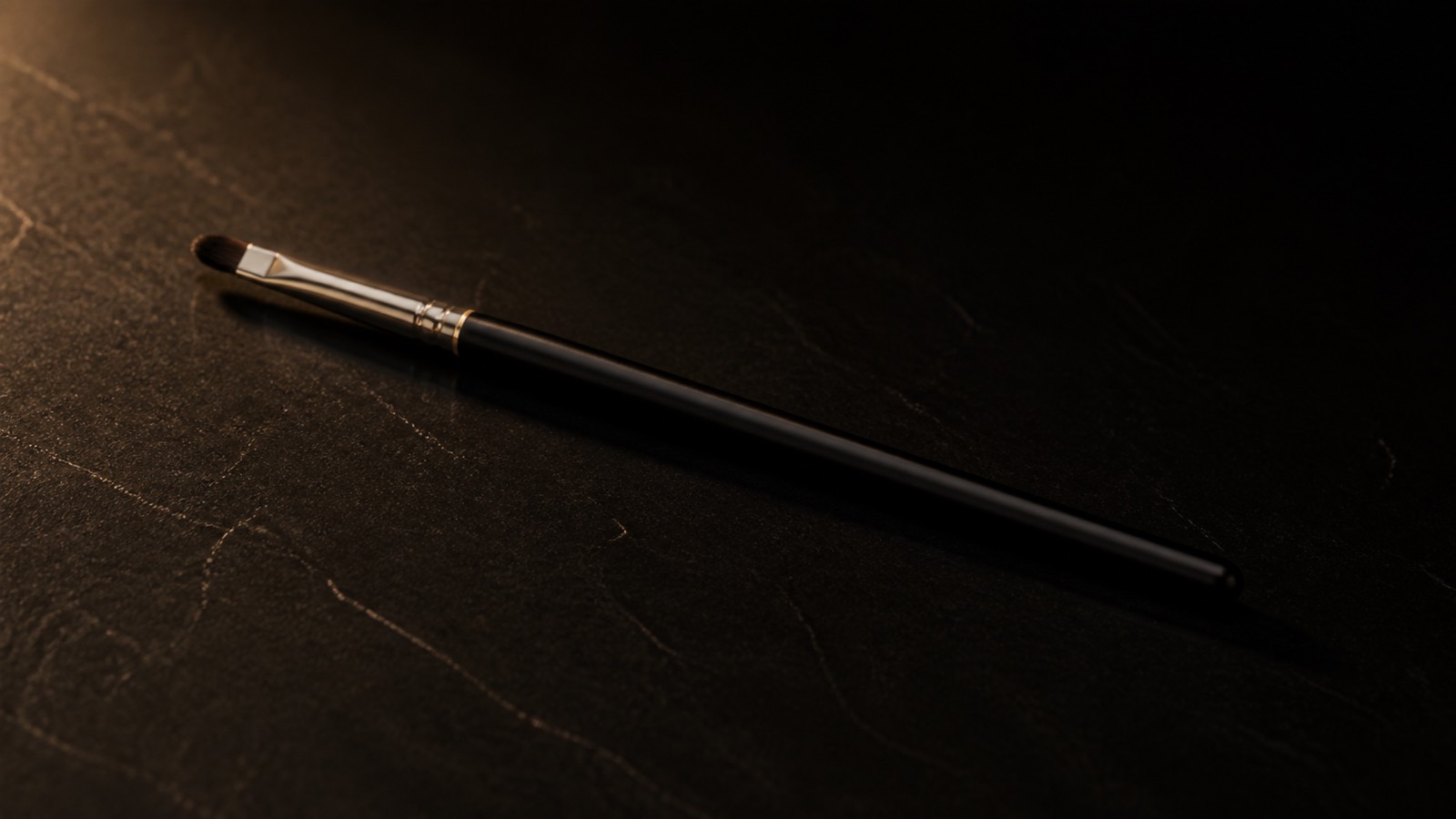 Pointed Lip Brush — seen in three-quarter view, low in a wide frame — under a single warm gold light on black.