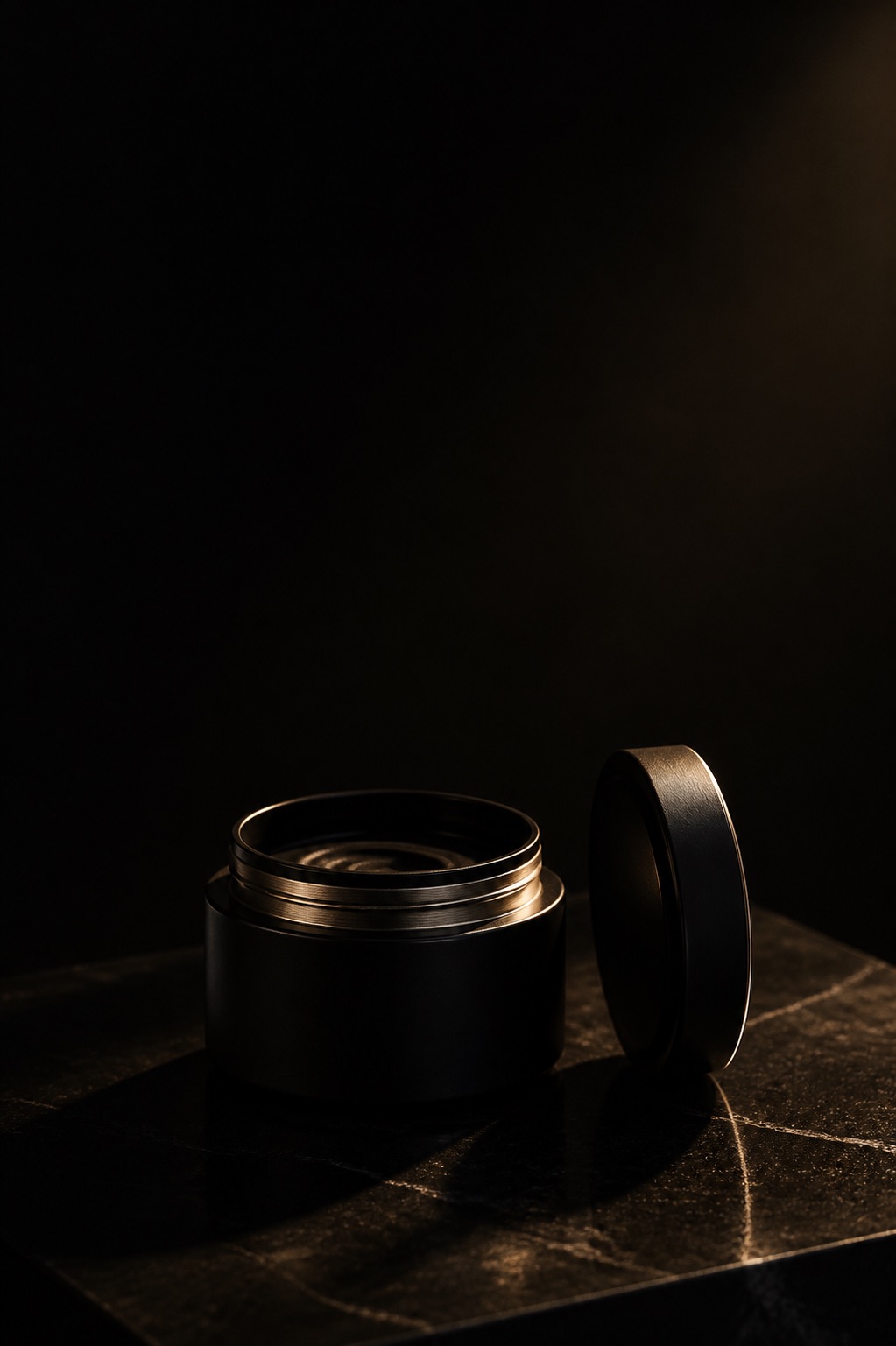 A matte black jar with its lid set down on its edge beside it, one gold thread of light along the open rim.