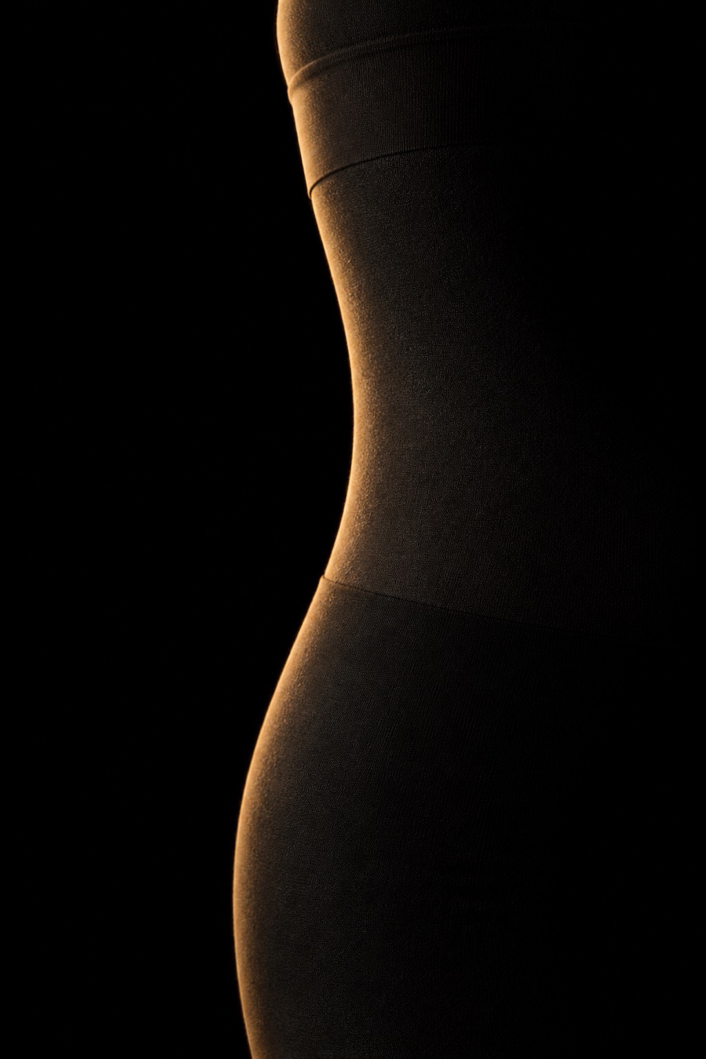 A seamless shaping garment on a featureless black form, its waist seam and the turn of the hip found by a single gold light out of the dark.
