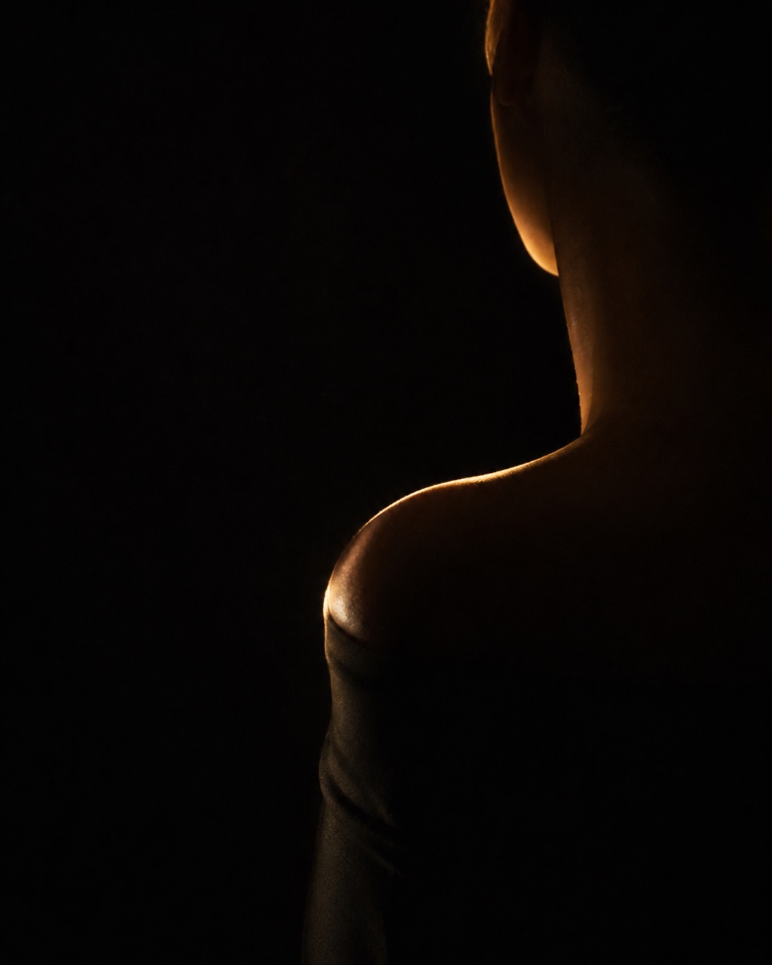 A shoulder and jawline turned away from the camera, traced by one gold rim light out of near-total darkness.
