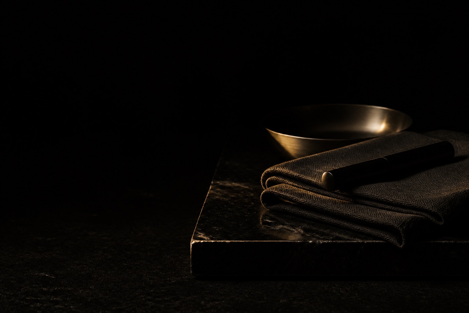 A dark worktable: black stone, folded linen, a steel dish and one unmarked object, gathered under a single low gold light.
