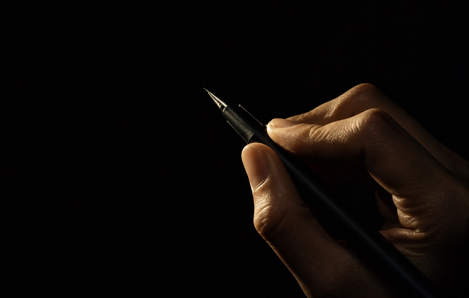 A hand at the angle of drawing, holding a fine black pen so one gold light catches the knuckles and the steel tip out of the dark.