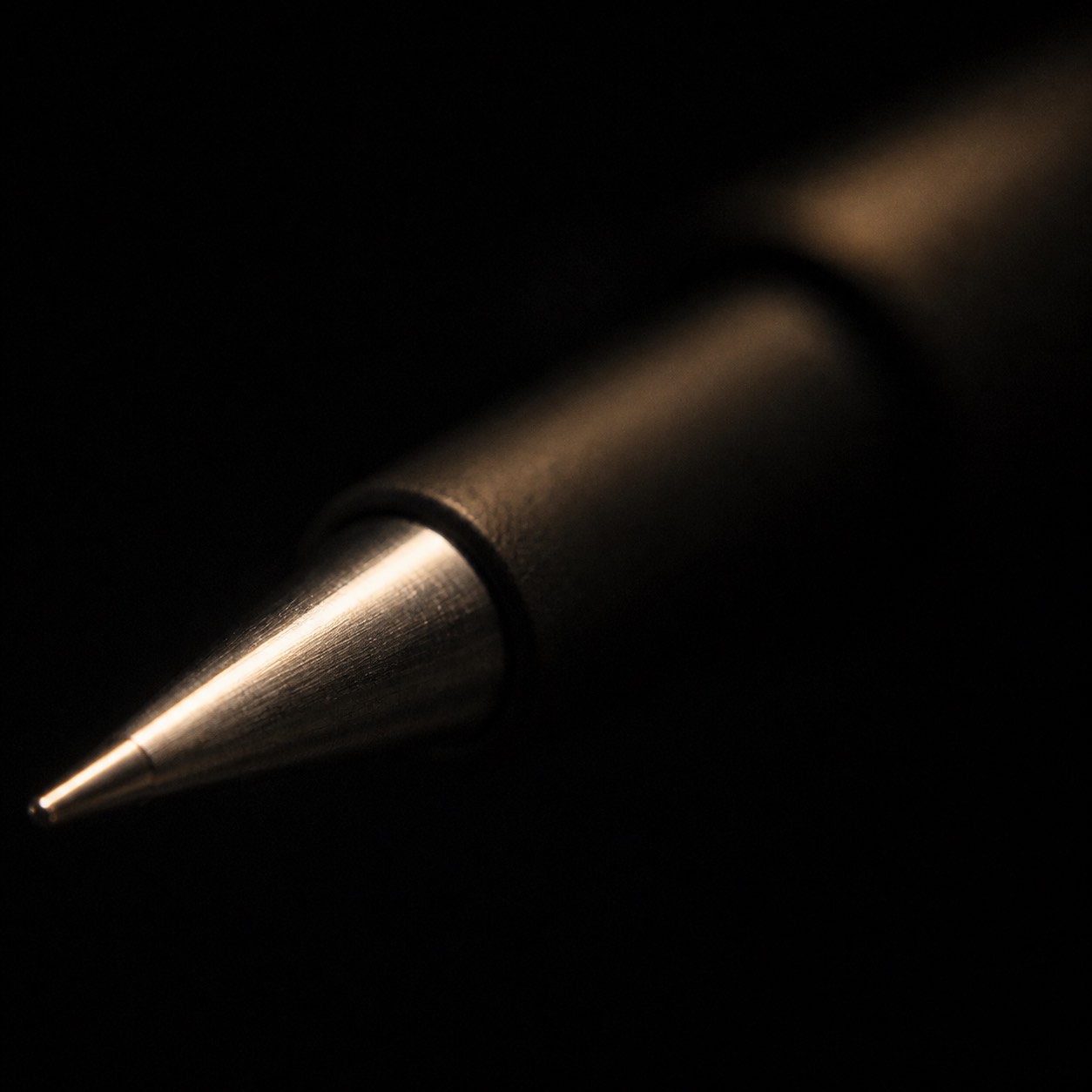 The tapered metal tip of a pen at close range, one gold light running along its edge and nothing else in the frame.