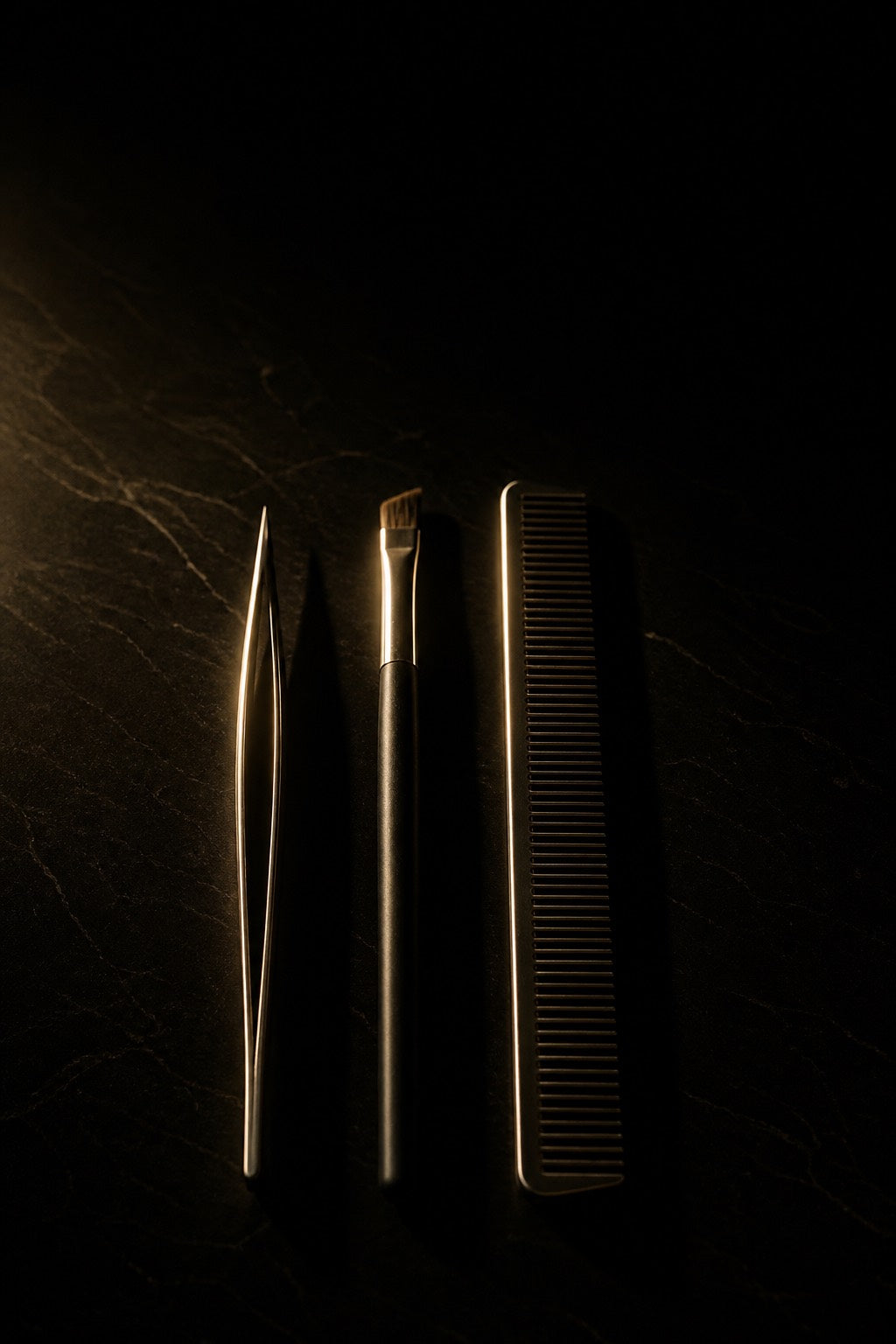 Three instruments laid in a row on black stone — a pointed tweezer, an angled brush and a fine comb — each found along one edge by a single gold light.