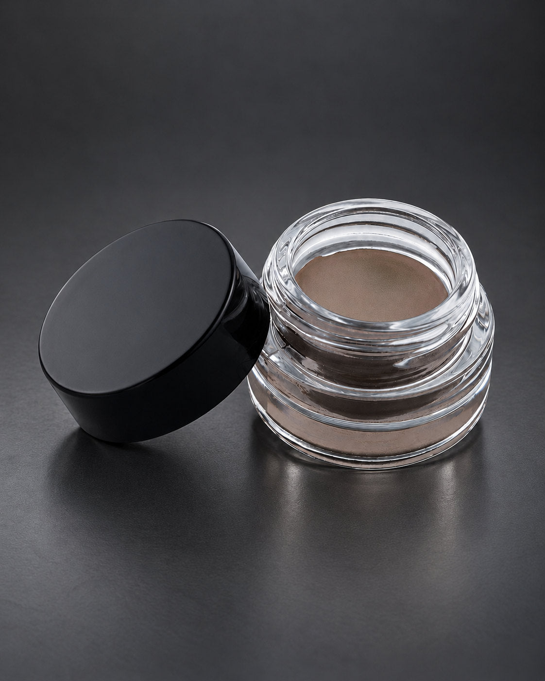 Brow Fixx in All in One: the clear glass jar open beside its black lid, on a charcoal studio surface. Digitally styled image.