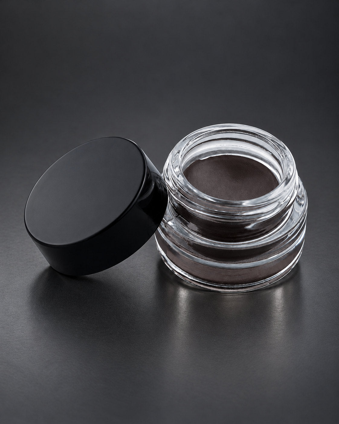 Brow Fixx in Black: the clear glass jar open beside its black lid, on a charcoal studio surface. Digitally styled image.