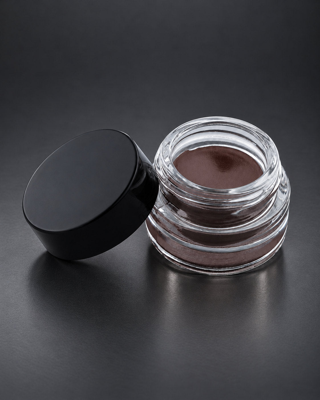 Brow Fixx in Dark Chocolate: the clear glass jar open beside its black lid, on a charcoal studio surface. Digitally styled image.