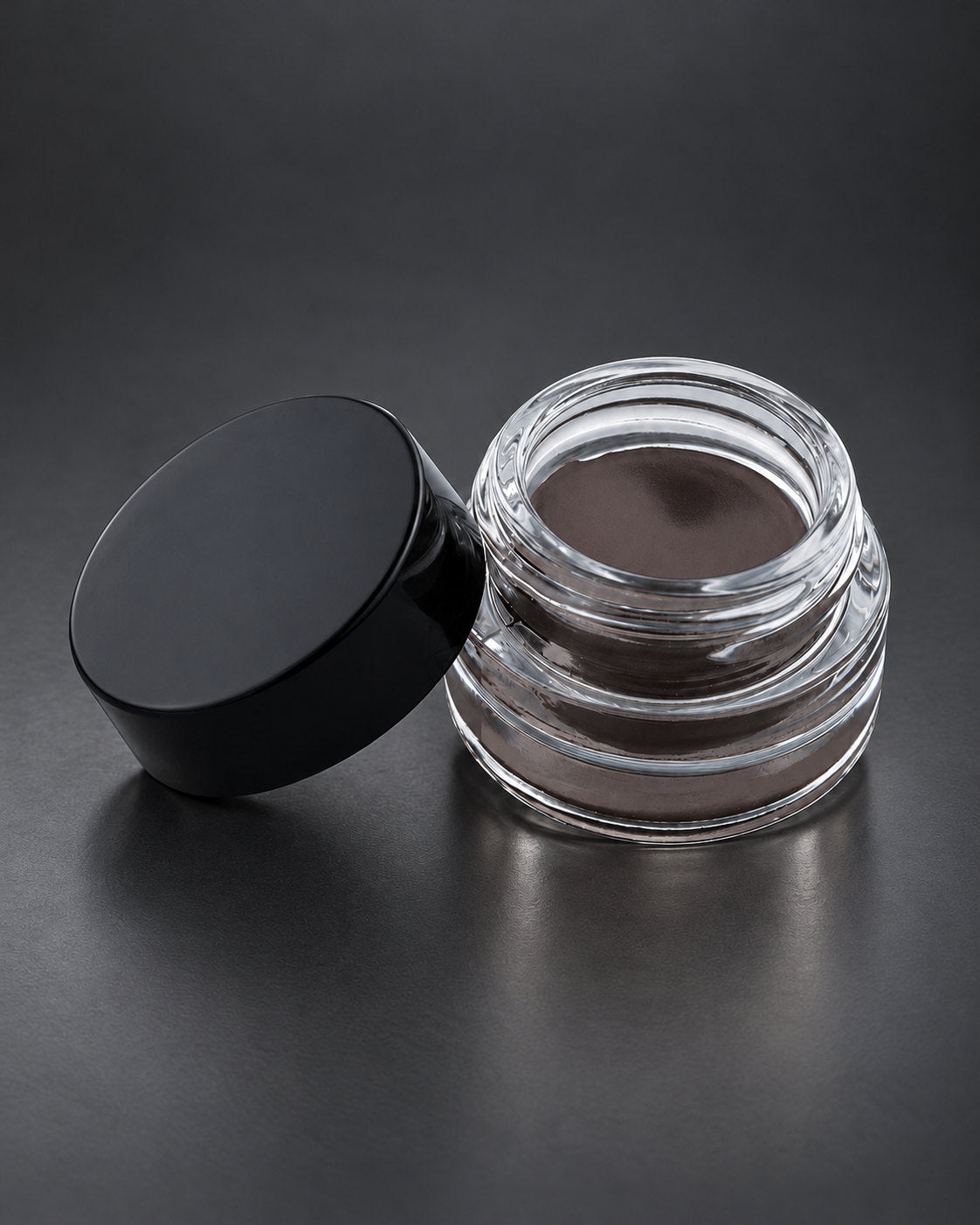 Brow Fixx in Midnight Sky: the clear glass jar open beside its black lid, on a charcoal studio surface. Digitally styled image.
