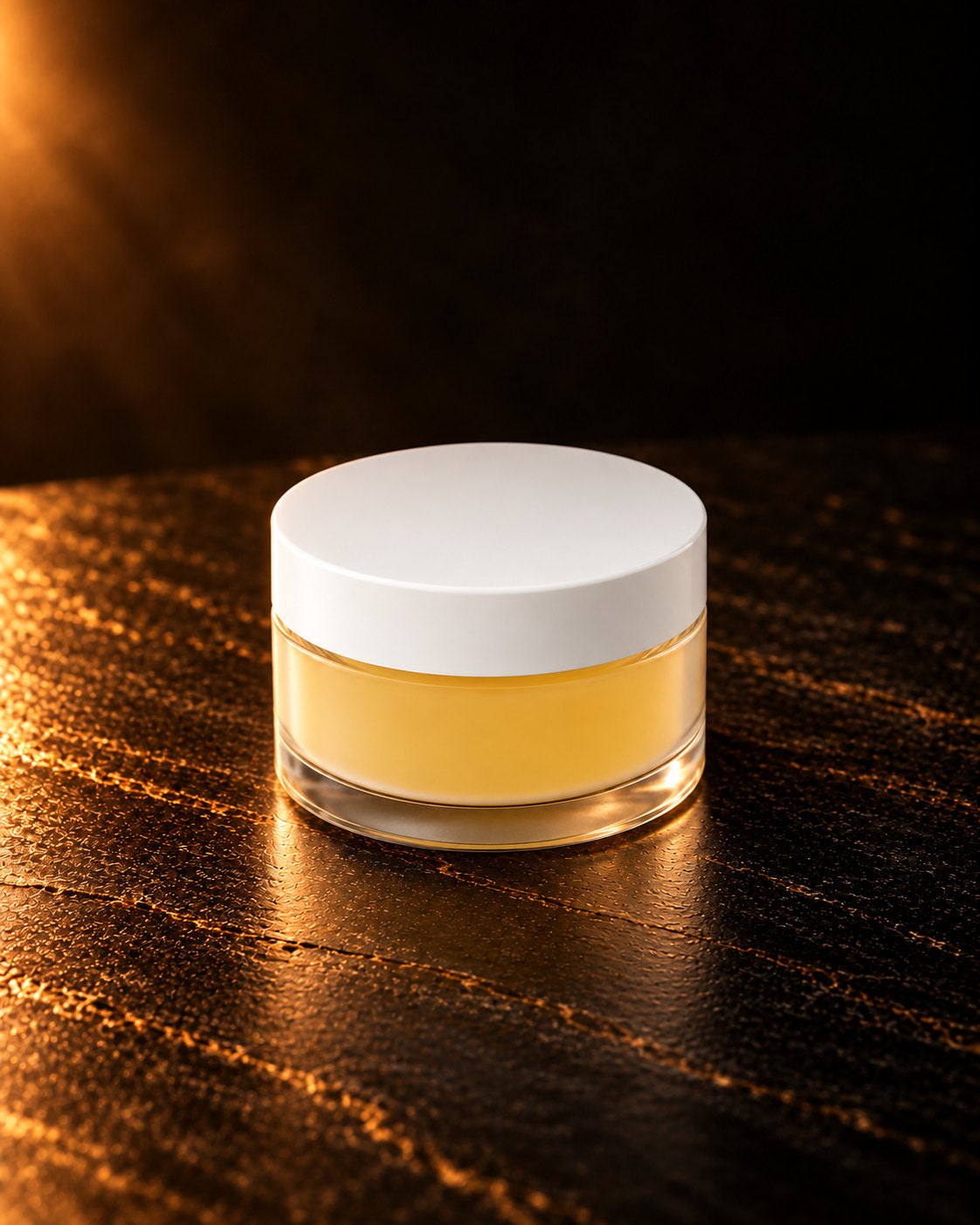 Lip Conditioner in Mango: a closed clear jar with a white lid and yellow Mango conditioner, on dark stone in warm side light. Digitally styled image.