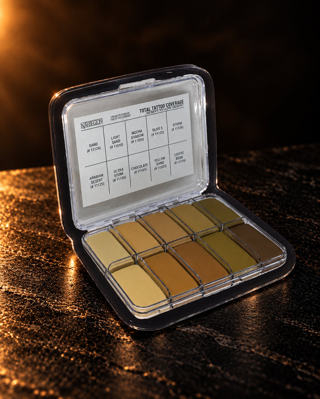 Total Tattoo Palette in Yellow Desert, open in its clear case with the shade chart in the lid and ten coverage pans, on dark stone. Digitally styled image.